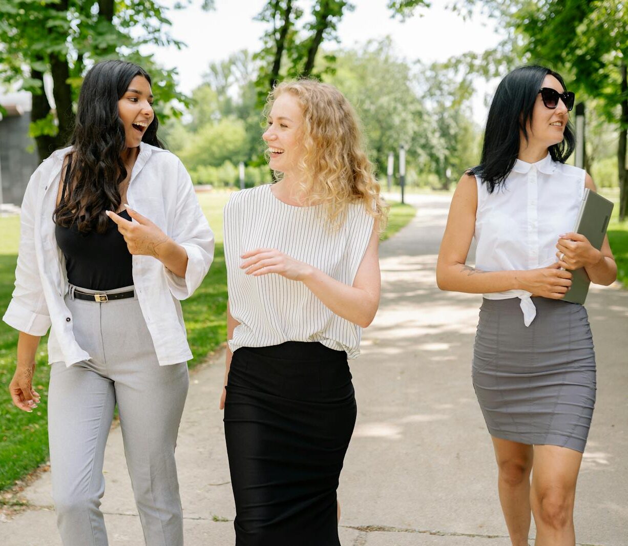 Three smiling businesswomen walk and converse in a sunny park setting, showcasing diversity and professional camaraderie.